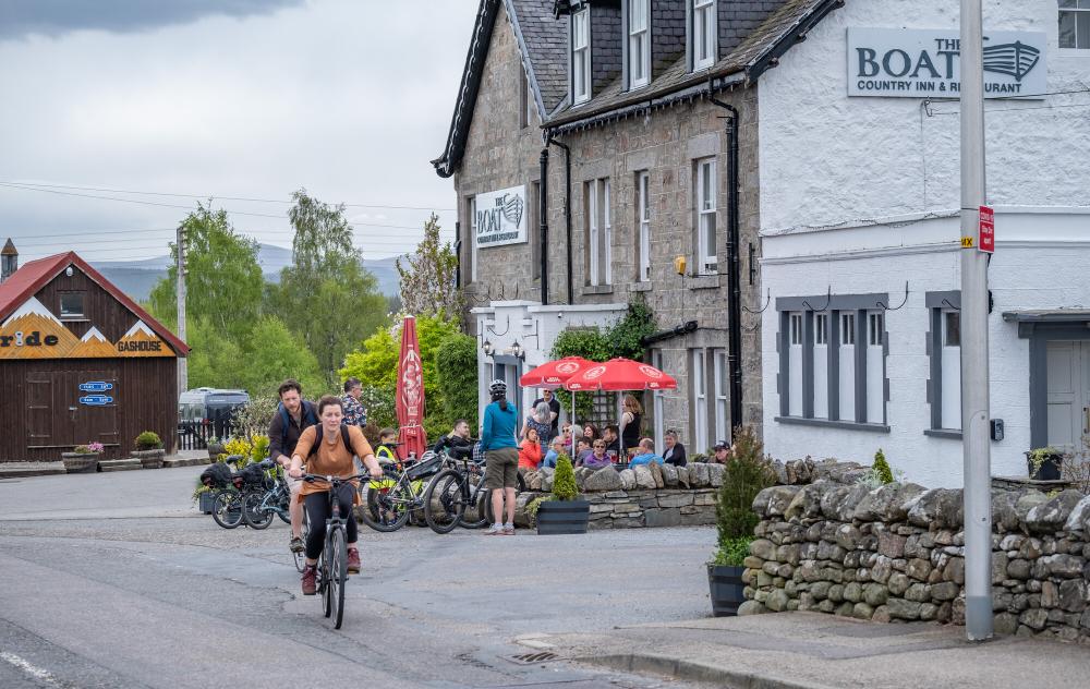 Two people cycling past the Boat of Garten hotel.