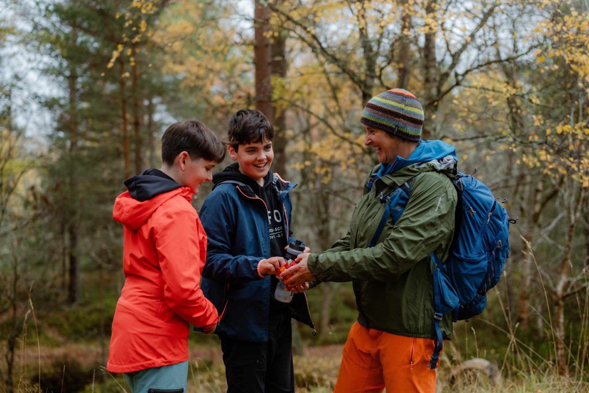 Three people share food in a forest, it is autumn and the leaves are turning. All three are smiling