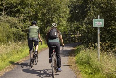 Two people cycle away on a path, it is sunny and both people are wearing helmets. There is grass on either ride and a sign for the path.