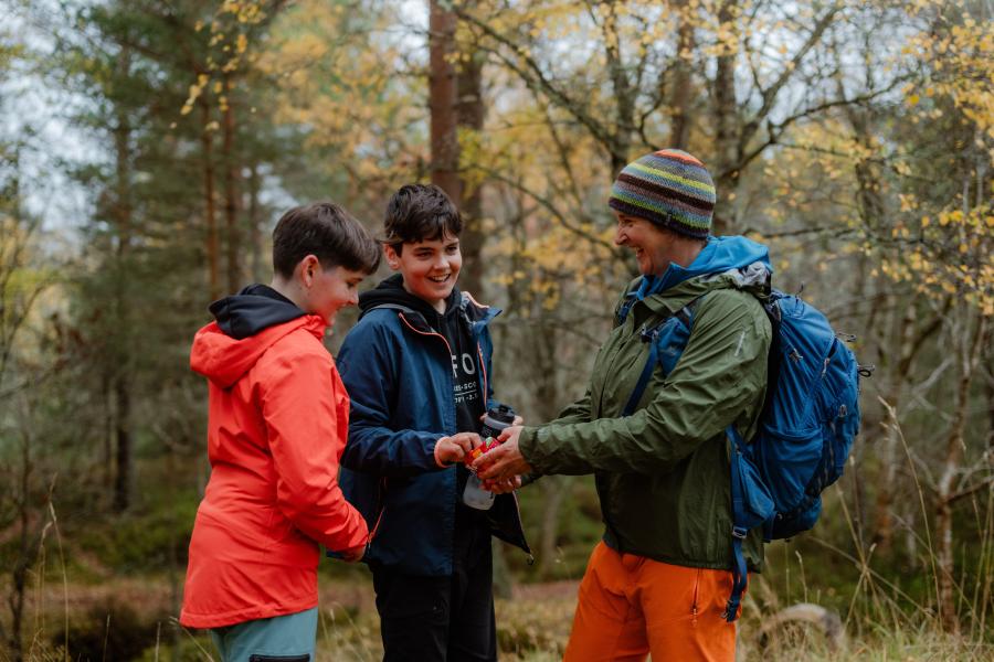 Three people share food in a forest, it is autumn and the leaves are turning. All three are smiling