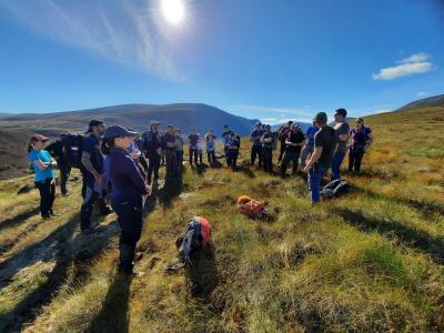 A large group of people congregate on a peat bog with mountains in the background on a bright, sunny day at Lynaberack
