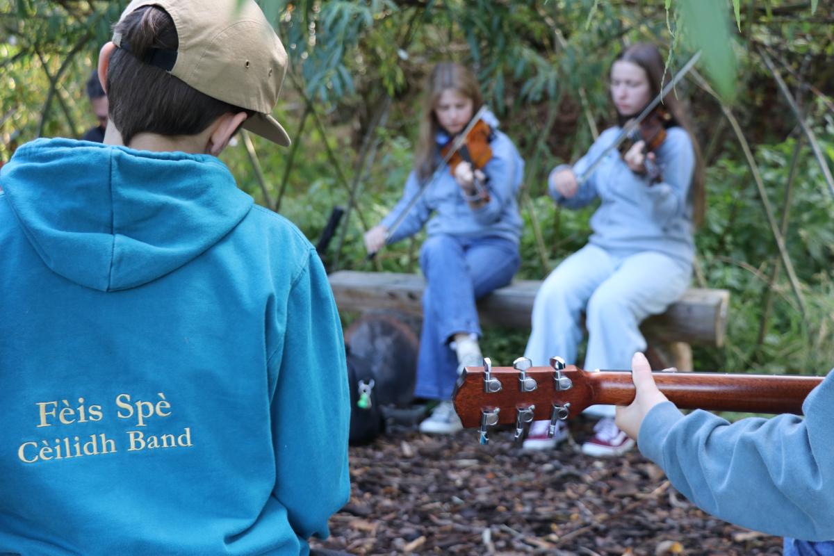 Four children sit in a tree-covered orchard practising playing on their instruments, including fiddle and guitar.
