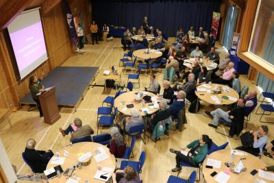 An audience sitting at tables in a hall listening to a speaker present in front of a screen.
