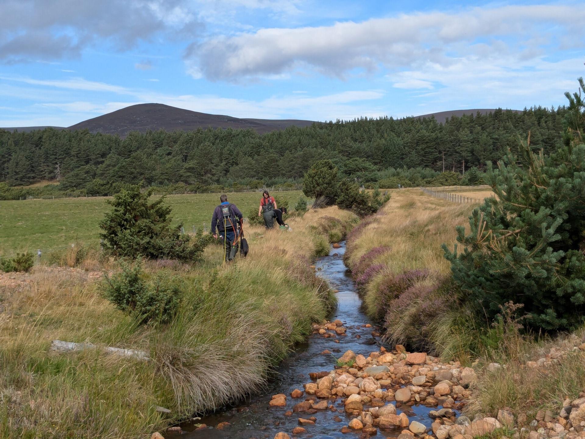 Three people on the banks of Slugain Burn with fields on either side.
