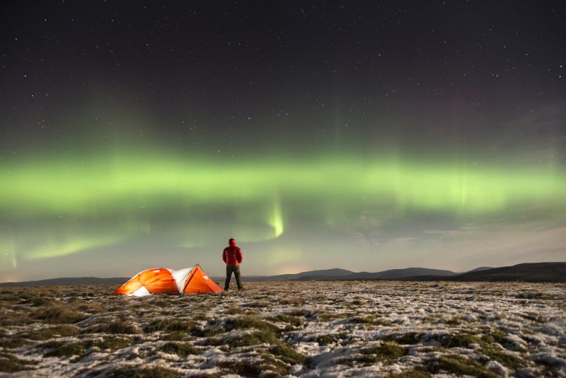 View of the Northern Lights/Aurora Borealis over the plateau of a mountain watched by a lone camper.