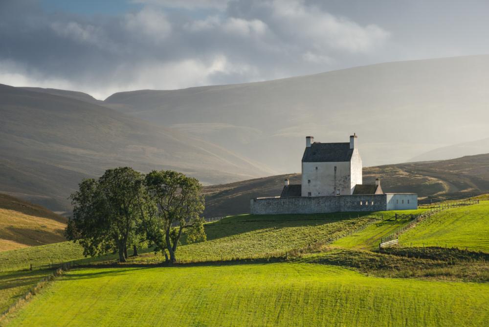 A fortified white walled castle with mountains in the background and fields in the foreground.
