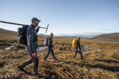 Three people walking across a peatland bog in hiking gear with backpacks, one is carrying a long tool over their shoulder