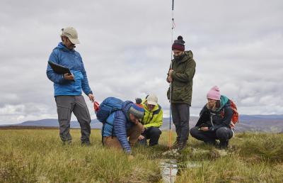 A man with a beard holds a long pole measuring the depth of a peat bog, whilst four colleagues closely examine the surrounding habitat
