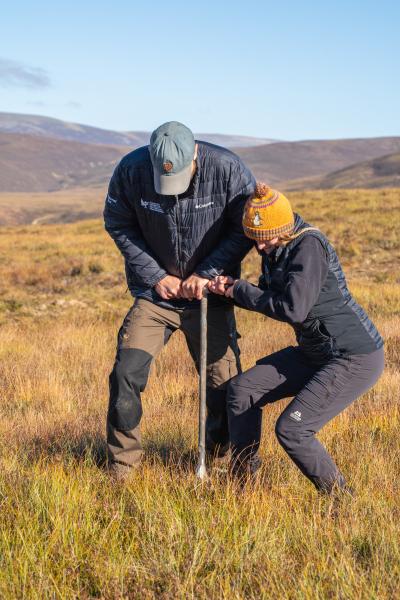 Two people on a peat bog turning a long vertical tool to bore it down into the peat beneath