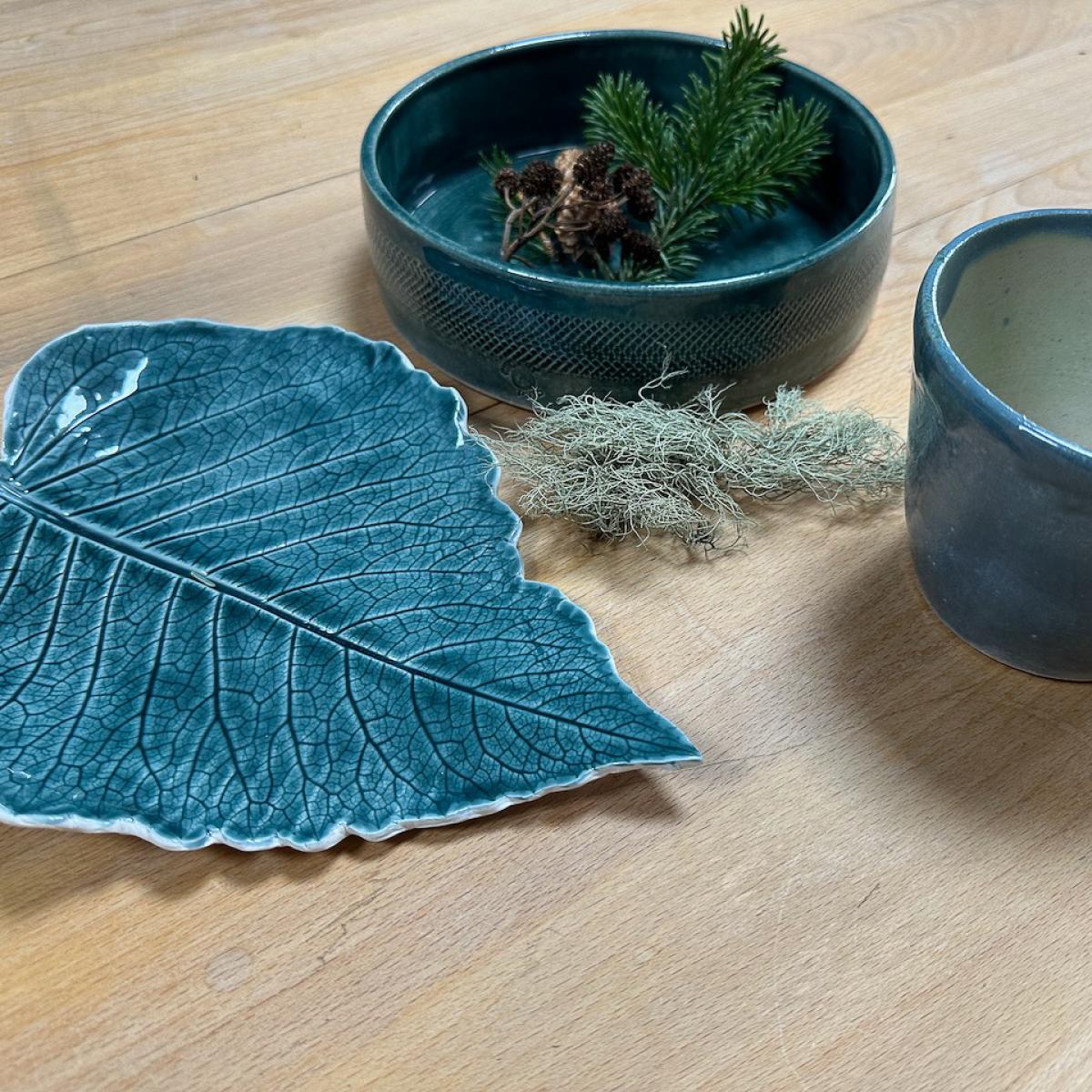 A blue, leaf-shaped dish sits on a wooden table alongside a blue bowl (with pine leaf in it) and a blue cup with white inside.