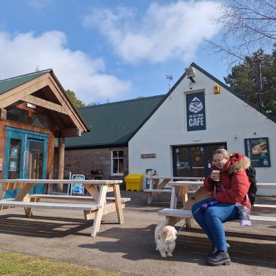 A visitor in a red jacket sits on a picnic bench outside Glenmore Visitor Centre with their white dog on a lead.
