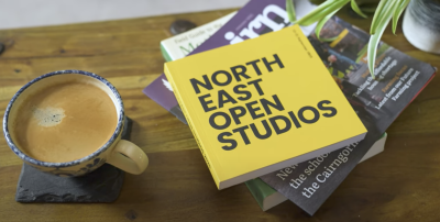Image of a cup of coffee on a slate coaster with a pile of books and brochures to the right. At the top is the new North East Open Studios brochure in bright yellow.