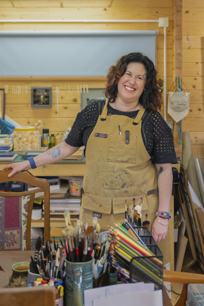 Photograph of artist Clare-Louise Battersby in her studio, surrounded by art materials including paint brushes, coloured pencils and canvases.
