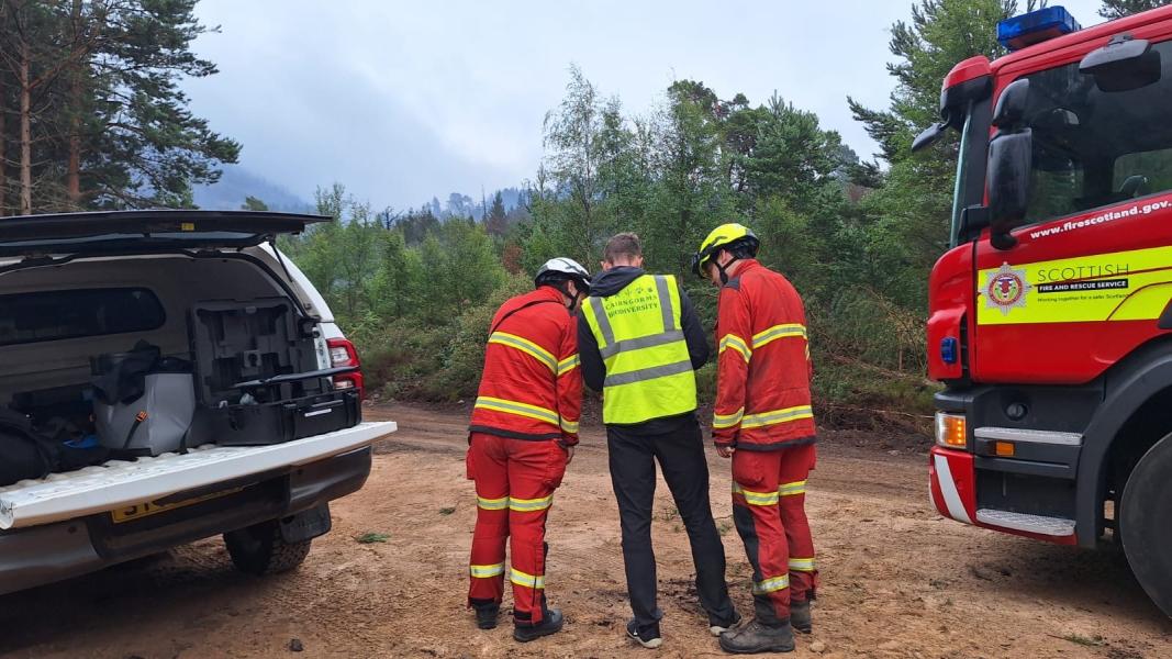A drone pilot shares video footage with two uniformed Scottish Fire and Rescue firefighters with a 4x4 and fire engine in the background