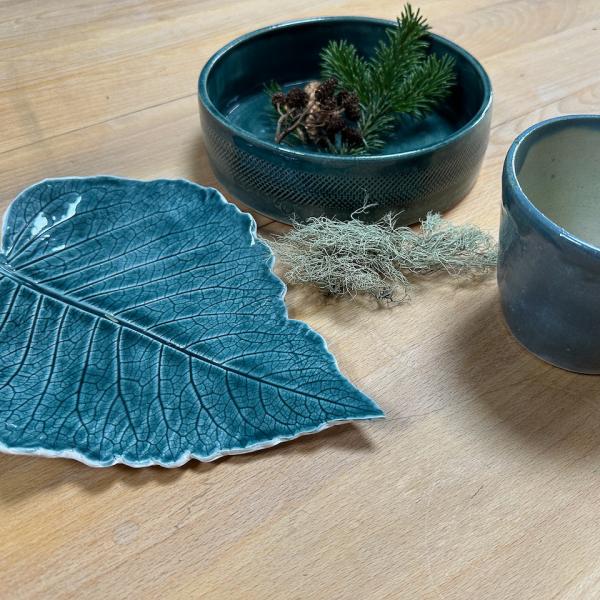 A blue, leaf-shaped dish sits on a wooden table alongside a blue bowl (with pine leaf in it) and a blue cup with white inside.