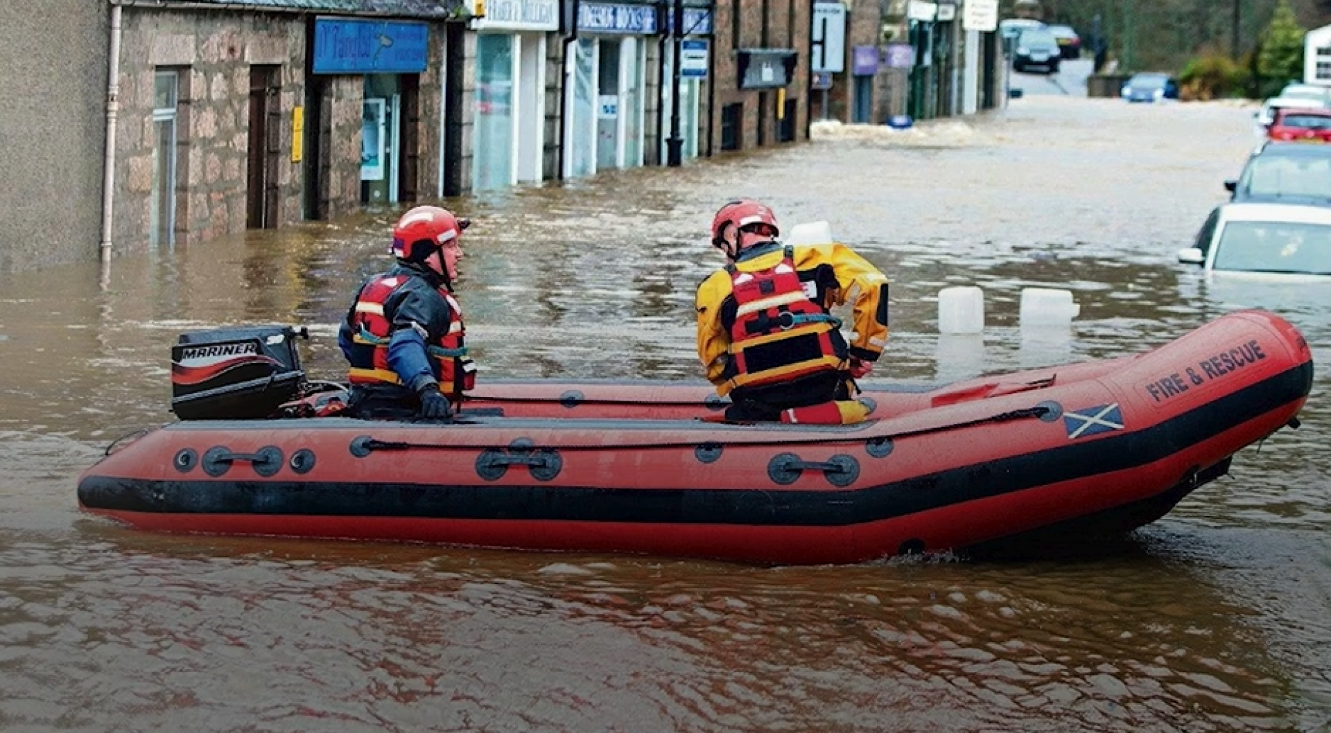 Two people sit in a small motorboat in a flooded high street