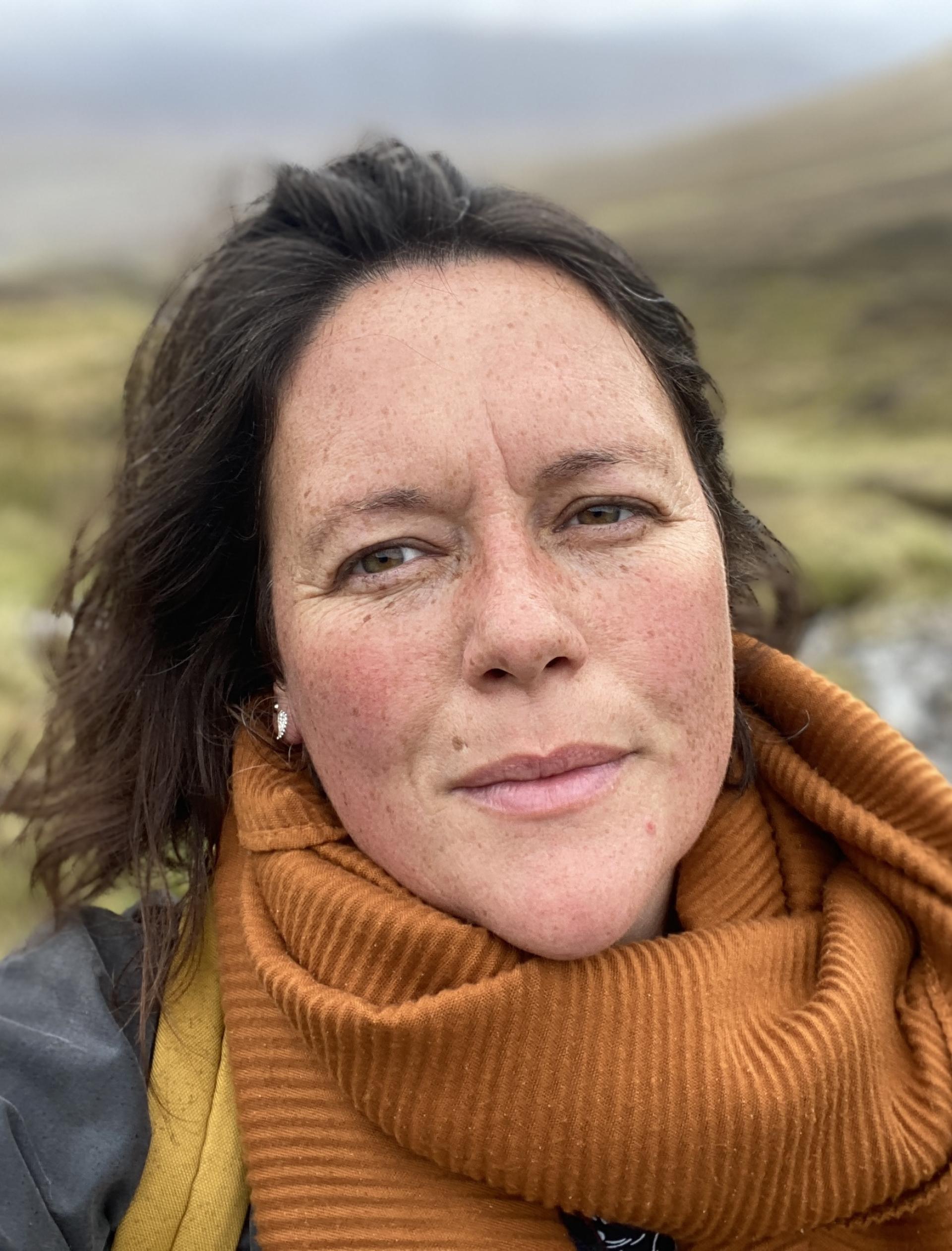 Close up of a woman's face, looking directly at the camera, with hills in the background.