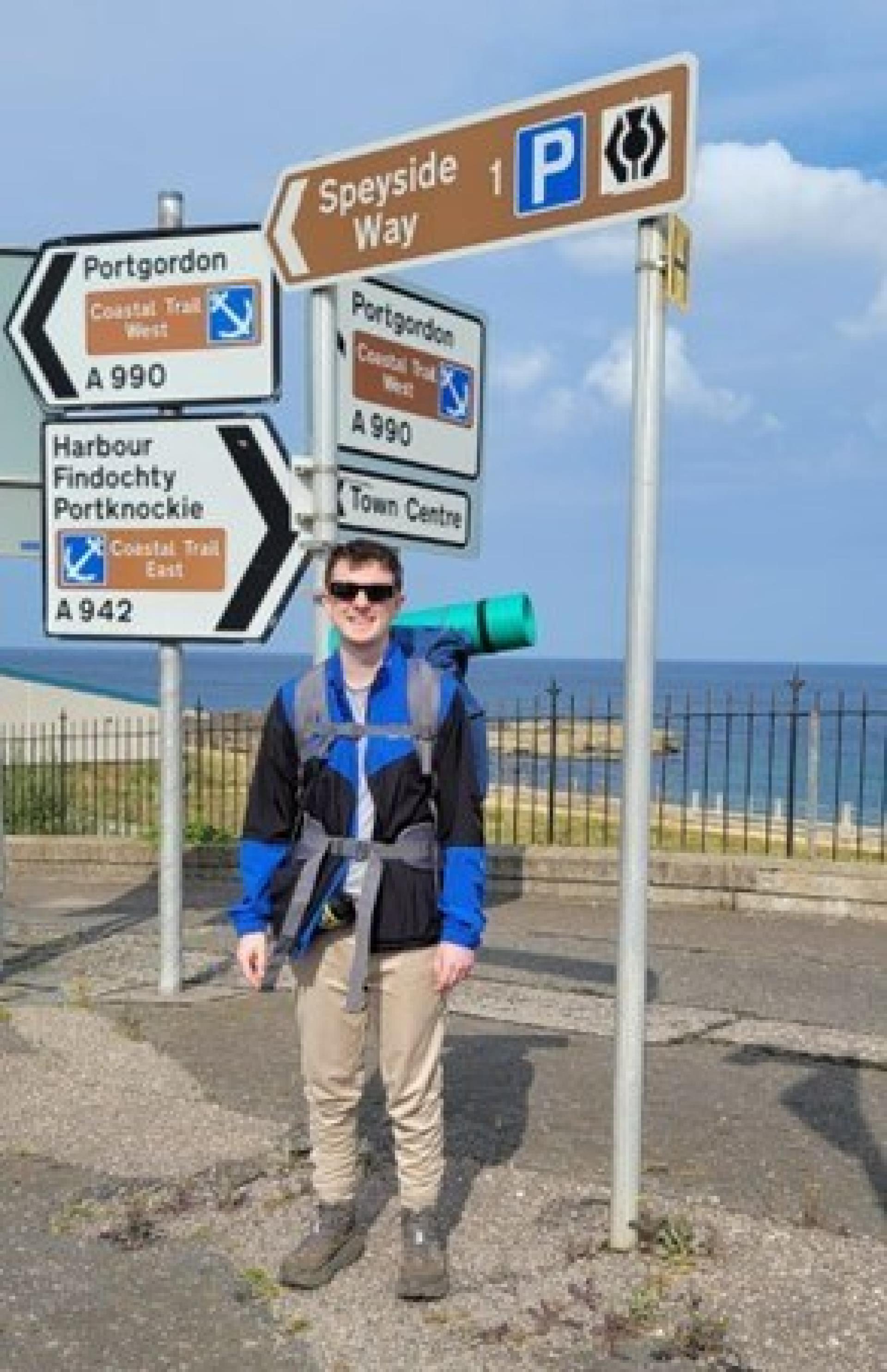 Shaun Patterson, wearing a backpack in front of a Speyside way sign in Buckie