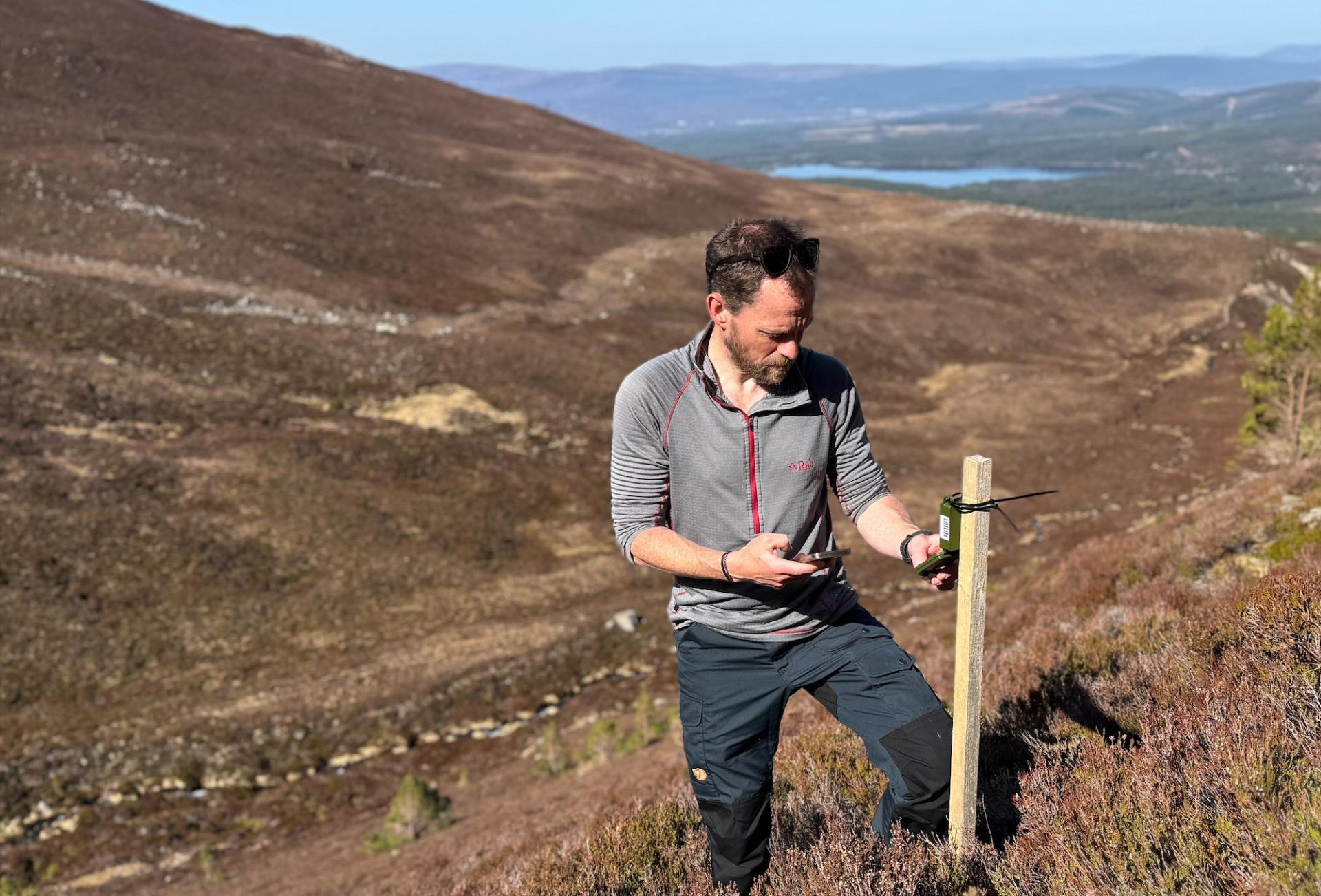 A person checking audio monitoring hardware in a mountain landscape.
