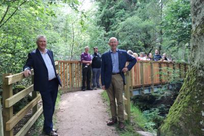 Two men stand in foreground of a bridge, with a group of people in the background.