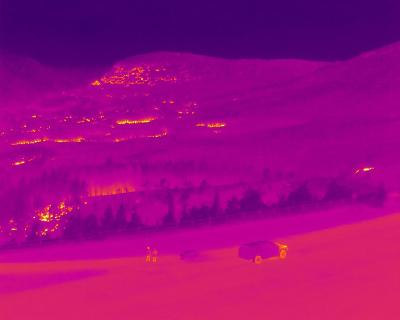 Thermal drone image showing two people and a vehicle in the foreground, with trees and flames in the background.