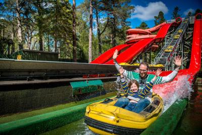 An adult sitting behind a child on a yellow and red water coaster ride with trees in background
