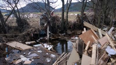 Sections of wood and buildings lie in a corner, with puddles of water in front of trees. Piles of debris lie in the field beyond.