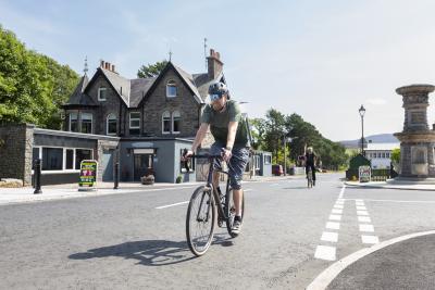 A person rides a bike on a clear road with Victorian-style houses behind, the sky is bright and blue