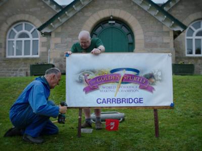 Two men installing sign for the Golden Spurtle World Porridge Making Championships in front of Carrbridge Village Hall.