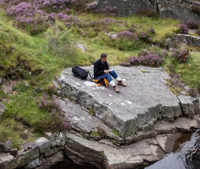 Alec Finlay, sitting on a large rock by the waterside in Glen Ey.