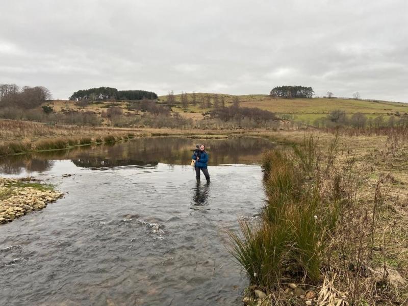 Image of Keilidh Ewan stood in the Beltie Burn on the River Dee holding a camera and microphone on her shoulder.