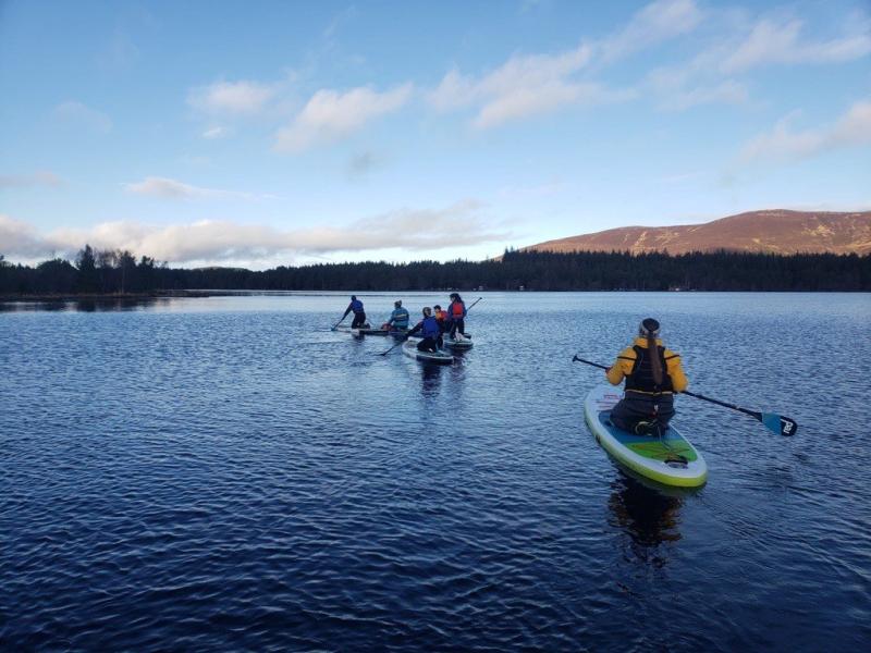 Six people kneeling on paddleboards on a loch with mountains in the background.