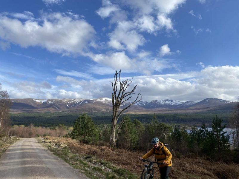 Image of Sarah Rathbone pushing her bike onto the trail with trees, a loch and a snowy mountain backdrop, after she stopped to take in the views.
