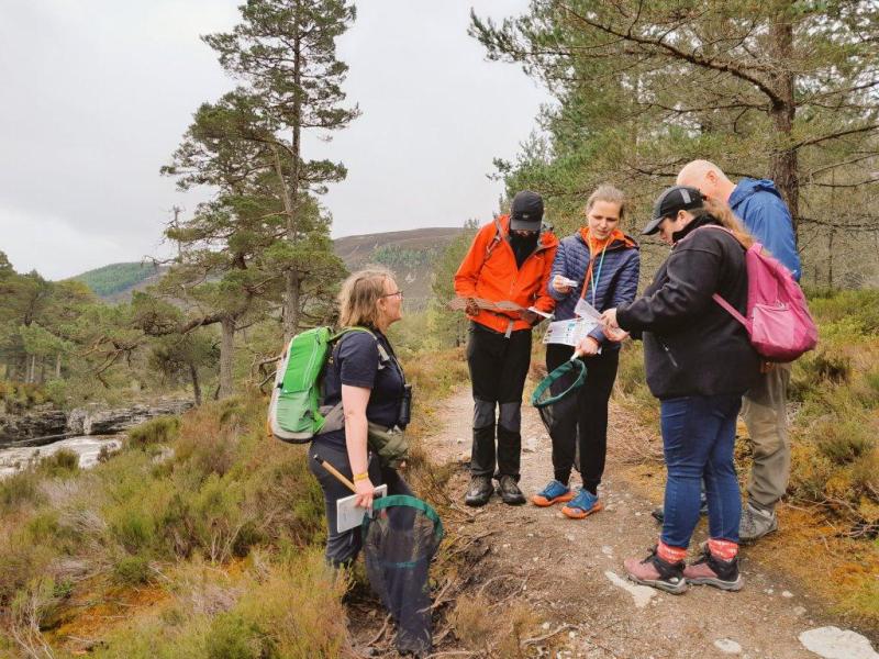 Image of five people, stood on a woodland trail above a river, two people are holding short handled nets, but all are looking at bee identification sheets to identify a bee temporarily caught in a in plastic tube at Mar Lodge.