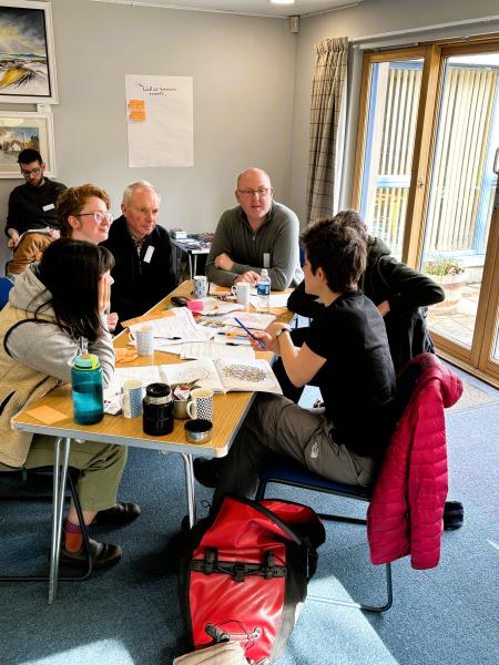 Five members of the Communities Fund panel plus a facilitator from Involve discuss options around a wooden tressle table in Boat Hall