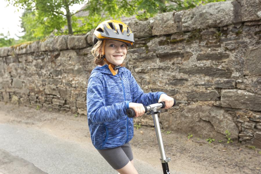 A young girl smiles at the camera while riding a scooter, she has a helmet on