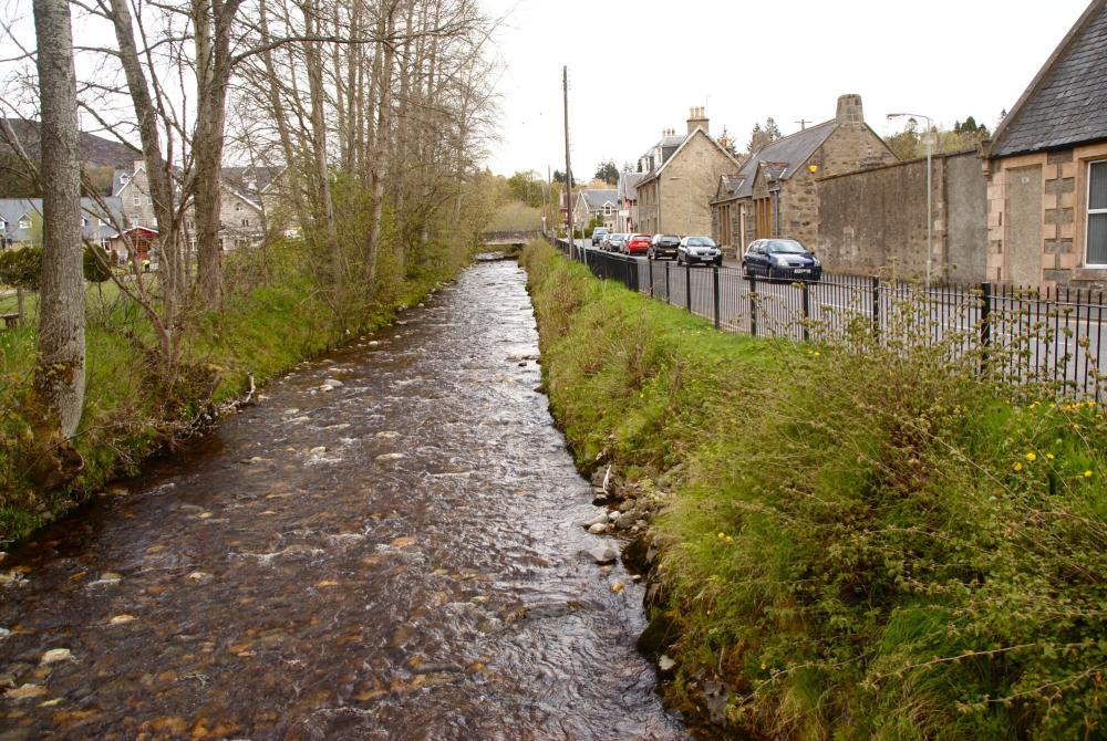 The Gynack burn flowing through Kingussie with trees on the left and a road on the right.