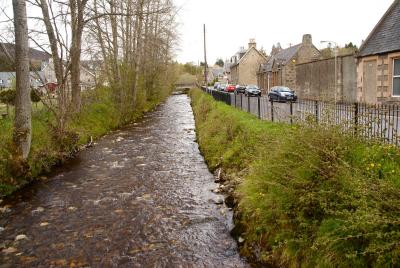 The Gynack burn flowing through Kingussie with trees on the left and a road on the right.