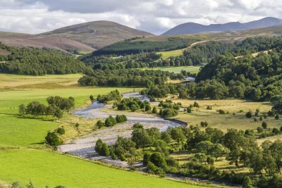 Winding river with fields to the left, trees to the right and mountains in the distance.