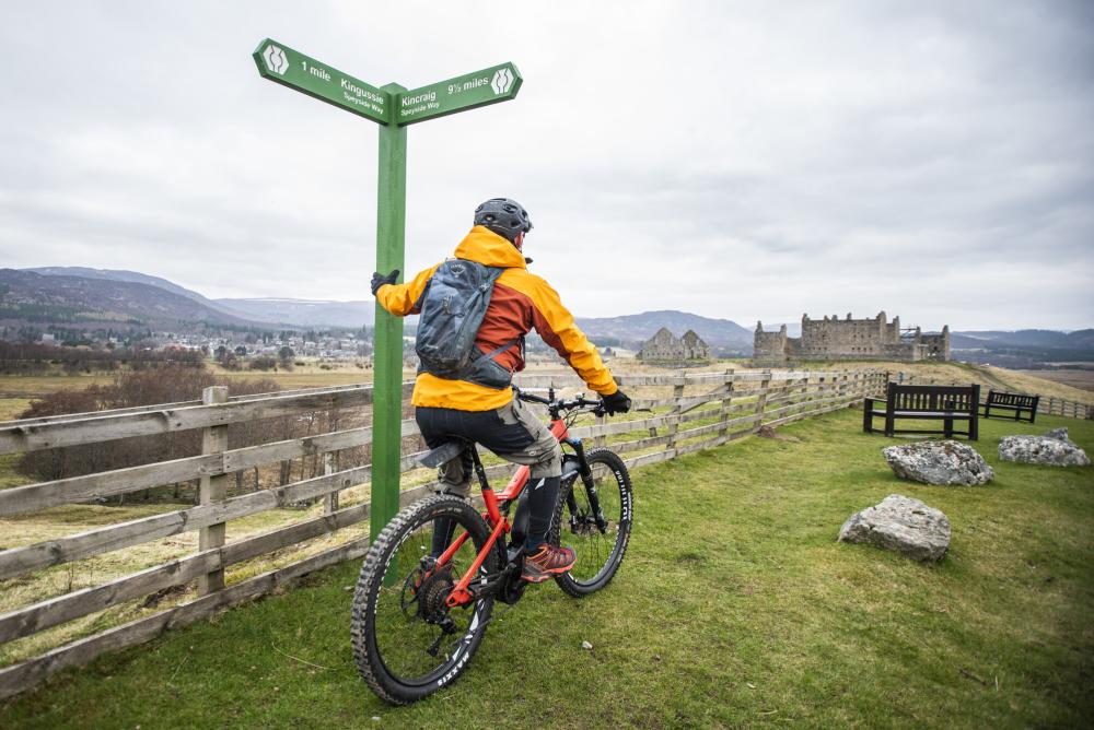 A cyclist leaning against a sign with Ruthven Barracks in the distance.