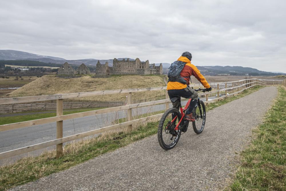 Person riding a bike on the Speyside Way path with Ruthven Barracks in the background.