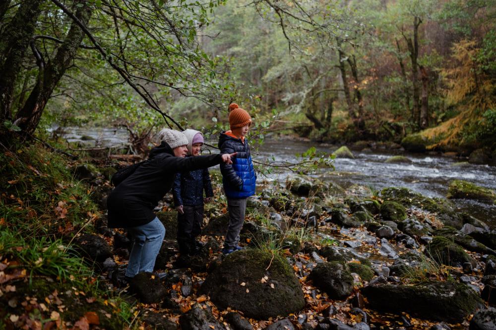 A family stand on the bank of a river as the mother points something out to the children. She is pointing out of frame and we can't see what she is pointing at.