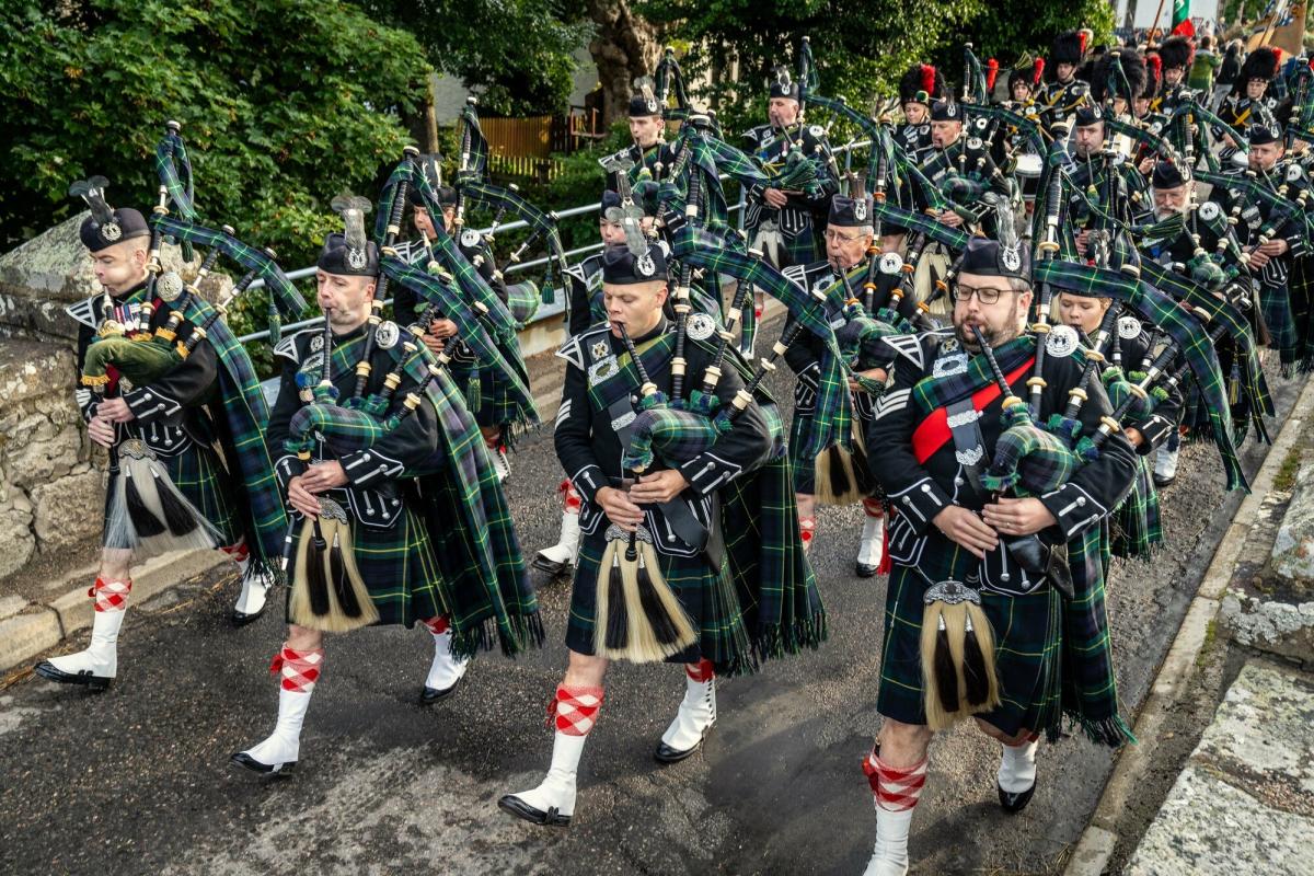 The Lonach Highlanders march across a bridge playing bagpipes.