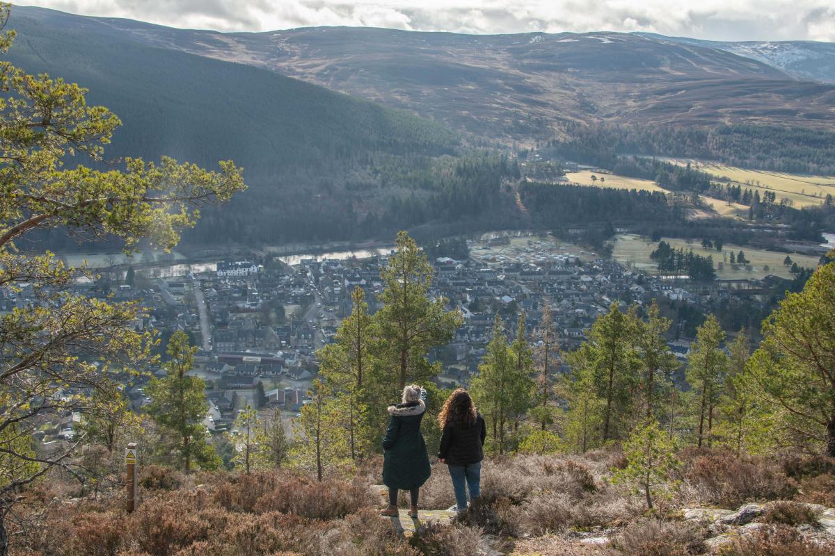 Two people in dark jackets looking out over trees and a town with hills in the background.