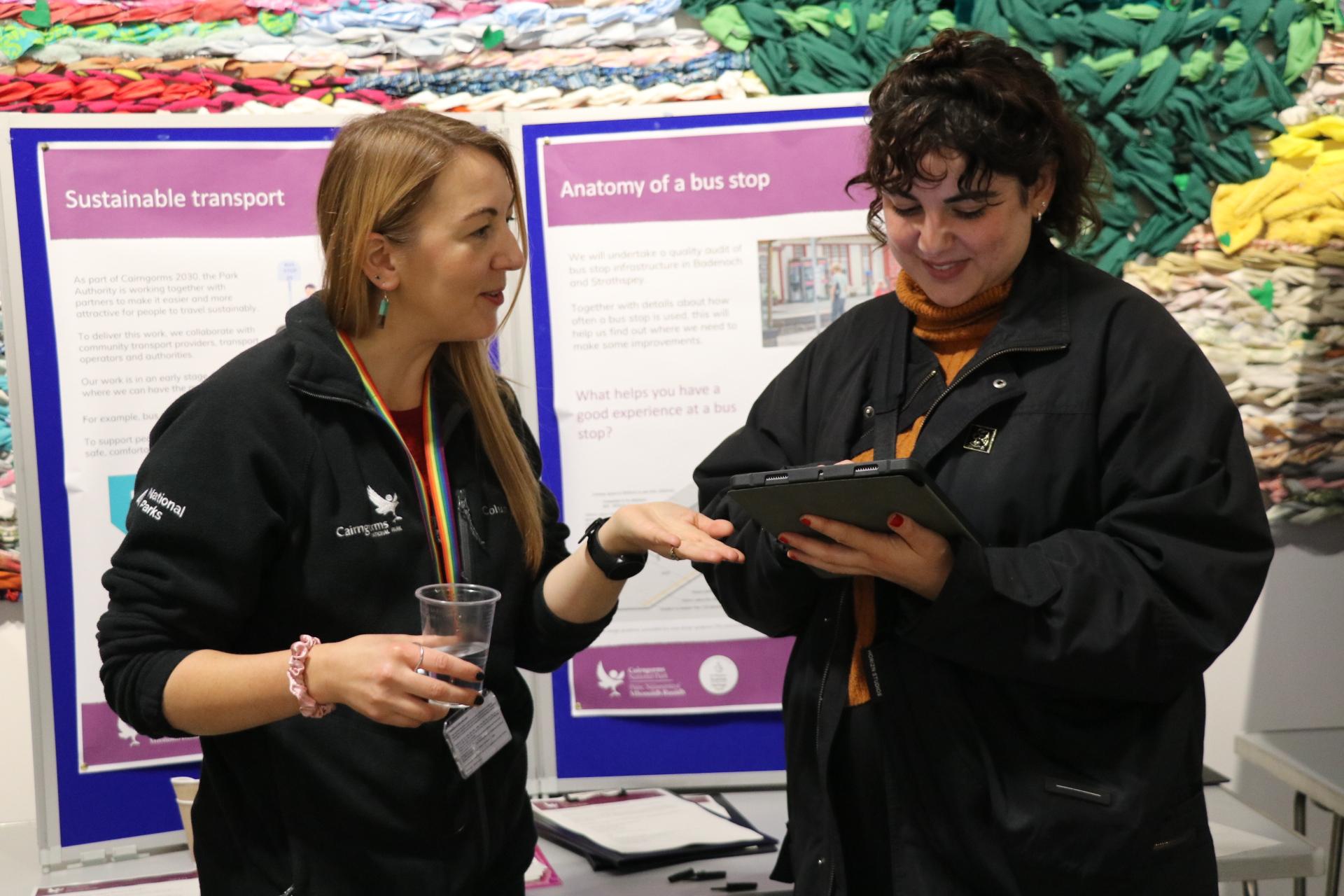 Two people chatting with information boards in background, one using a tablet.