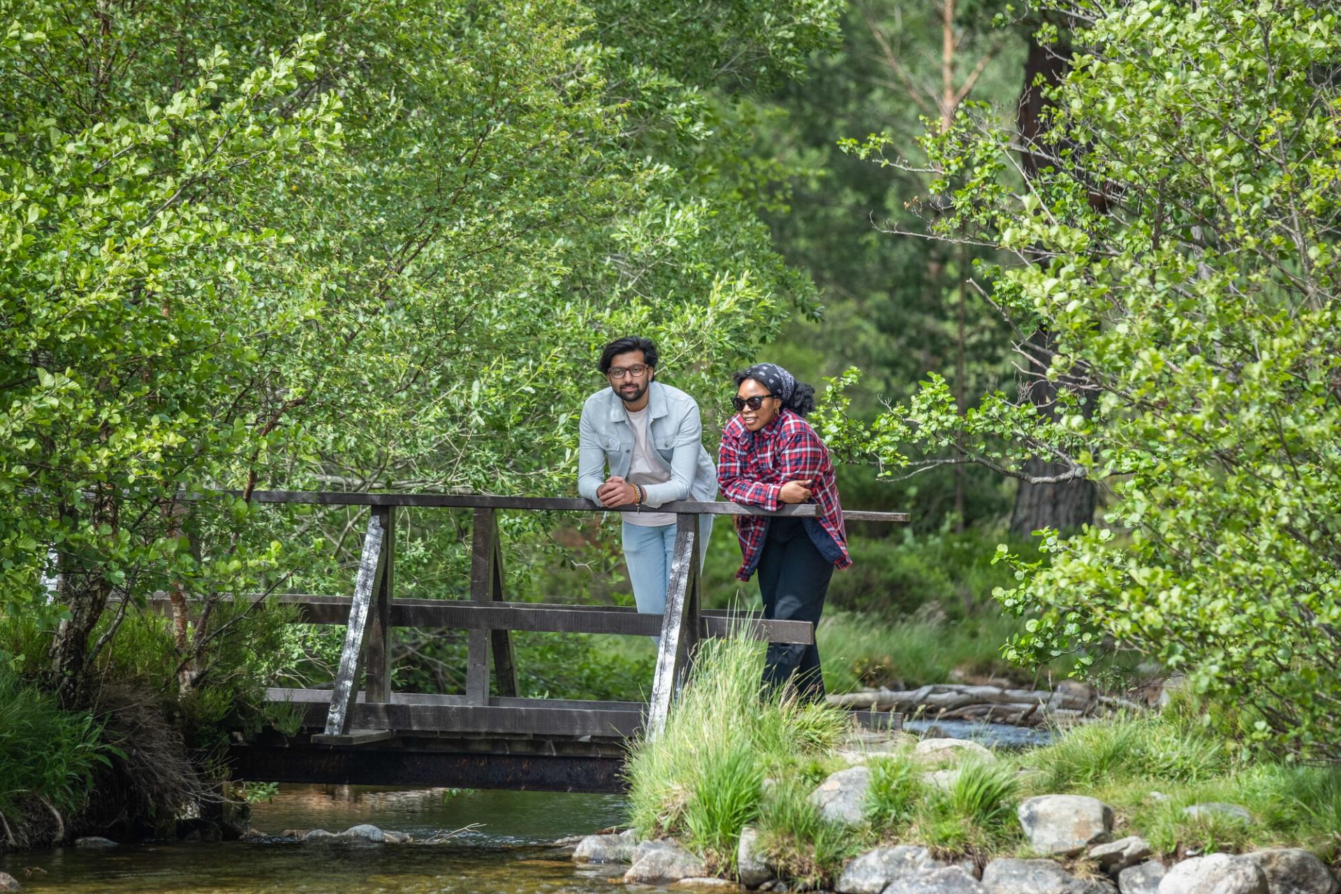 2 people leaning on a small bridge over a stream of water surrounded by trees