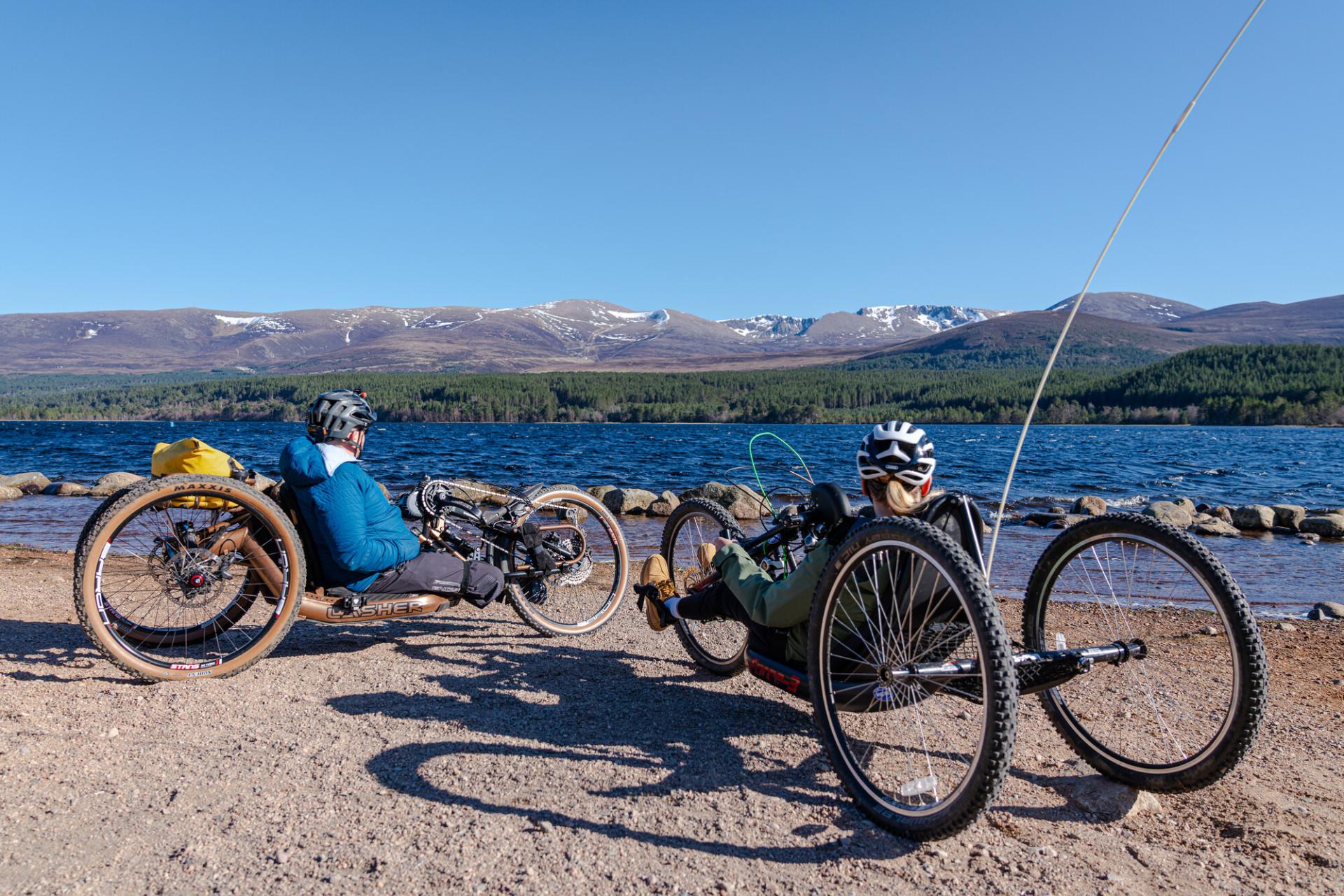 People using adaptive bikes at Loch Morlich beach with loch and mountains in the background.