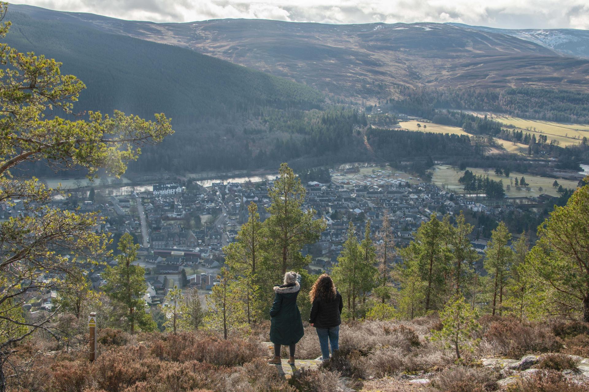 Two people in dark jackets looking out over trees and a town with hills in the background.