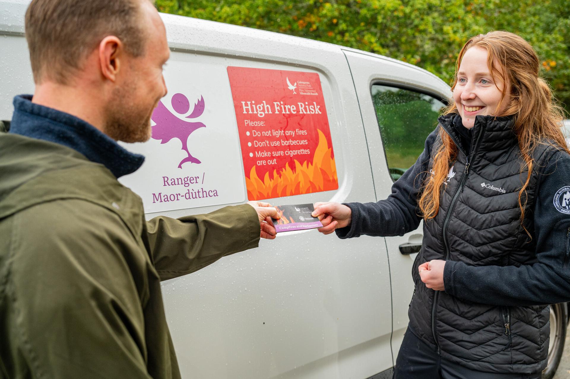 Seasonal Ranger hands a fire info leaflet to a member of the public, next to a van stating 'High Fire Risk'.
