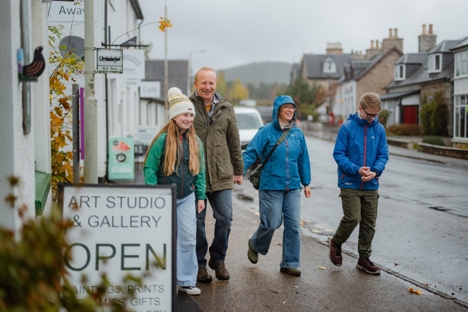 A family of four walk down a street past shop signs that say 'Open' and 'Take Away' and a bus stop. The weather is wet and the sky grey, but the family are smiling.